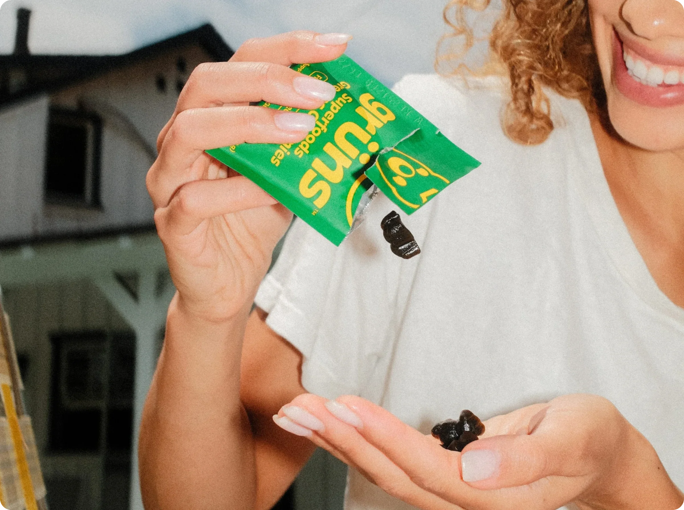 A smiling woman in a white t-shirt pours a dark gummy from a green Grüns pouch into her other hand, outdoors in front of a house.